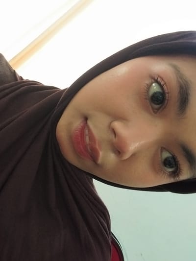 A close-up, angled selfie shows a young woman's face, tilted downwards. She is wearing a dark brown hijab that covers her hair and neck. Her makeup is visible, with mascara accentuating her eyelashes and red lipstick on her lips. The background appears to be an indoor setting, with a bright, white surface above and to the left, possibly a wall or ceiling with a light source. The lighting creates a slight glow on her skin. No specific location in Watampone, Indonesia, can be identified from the visual information provided.