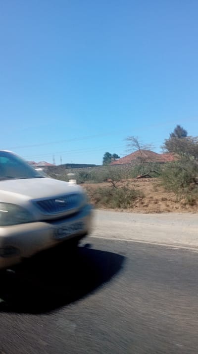 A blurry image shows a silver-beige car moving quickly along a dark asphalt road. The sky is a clear, bright blue. A grassy verge with small bushes and trees runs alongside the road. In the background, a house with a red tile roof can be seen. Power lines are visible above the bushes. The car's shadow is large and dark, stretching across the road.