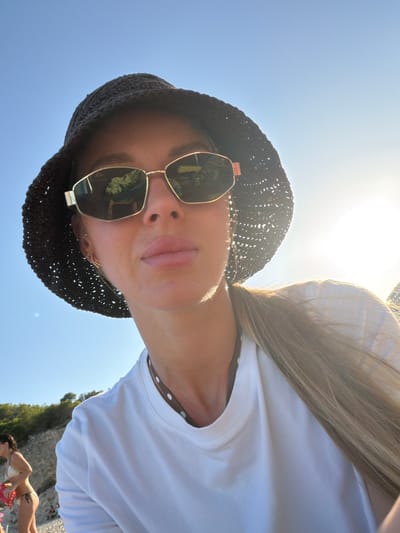 This media file is a first-person perspective selfie taken outdoors, likely at a beach or coastal area. The subject is a woman wearing a dark, crocheted sun hat and stylish sunglasses with gold frames. The sunglasses reflect a scene that appears to be outdoors, possibly with foliage and some red objects. Her hair, which is long and blonde, is visible and catches the sunlight. She is wearing a white t-shirt with a subtle polka dot trim visible at the neckline.
The sky is a clear, bright blue, and the sun is high and strong, indicating it is daytime, likely midday, and the weather is sunny and warm. The angle of the photo suggests the subject is looking up towards the sky or taking the picture from a low vantage point.
In the background, there are hints of a rocky cliff face covered in greenery, characteristic of a coastal landscape. Further in the distance, other people can be seen in swimwear, suggesting a beach setting. The overall mood appears relaxed and summery. No visible text is present in the image. The orientation of the photo is portrait, rotated 90 degrees counter-clockwise.