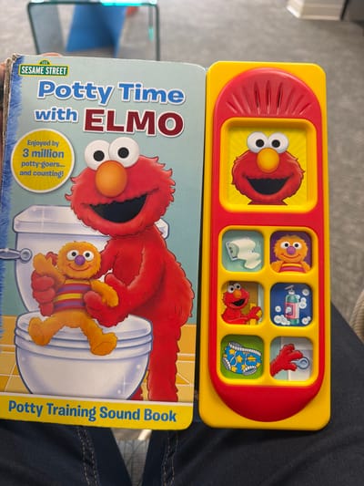 A "Potty Time with Elmo" sound book is displayed next to a yellow and red Elmo-themed electronic toy. The sound book features an illustration of Elmo and a character named "Grover" sitting on a toilet. The electronic toy has a red curved top and displays six small squares with images of Elmo characters and related objects, such as toilet paper and soap. The book's cover also advertises "Sesame Street" and mentions that it is "enjoyed by 3 million potty-goers... and counting!". The scene appears to be indoors, possibly in a home or classroom environment, but the specific location in Chesapeake, United States of America cannot be determined from the image.