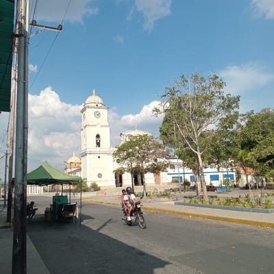 The image shows a street scene in Duaca, Venezuela. A light-yellow clock tower with a dome and a visible clock face stands prominently in the background, partially obscured by a tree. The sky is blue with scattered clouds. In the foreground, a motorcycle with two riders is on the street. The road has a light-colored curb. A building with white walls and blue-trimmed windows is visible adjacent to the tower. An outdoor vending stall with a green awning stands near the bottom of the frame. A person is sitting on the ground near the stall. Utility poles and wires are present in the lower left corner.