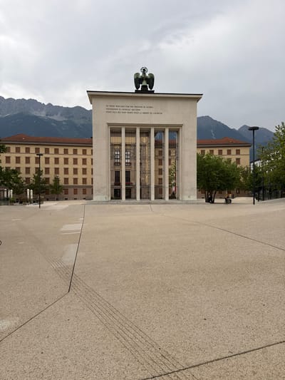 The image shows a memorial building with an eagle statue on top, situated in a plaza. The building is made of light-colored stone and features large, ornate windows with metal grills. Inscribed on the facade is text in English, Russian, and French, reading "TO THOSE WHO DIED FOR THE FREEDOM OF AUSTRIA," "ПОГИБШИМ ЗА СВОБОДУ АВСТРИИ," and "POUR CEUX QUI SONT MORTS POUR LA LIBERTÉ DE L'AUTRICHE." Behind the memorial, a pale yellow building with rows of brown-windowed facades is visible, with trees and a lamppost in front of it. The background is dominated by imposing, mist-covered mountains, suggesting a mountainous region. The plaza is paved with light-colored stone, with distinct lines and geometric patterns. The sky is overcast, indicating a cloudy or possibly rainy day. A few people are seated in the distance, near the trees, adding a sense of scale and human presence to the scene. The overall atmosphere is somber and reflective, befitting a memorial site. The location is identified as Innsbruck, Austria, which aligns with the mountainous landscape.