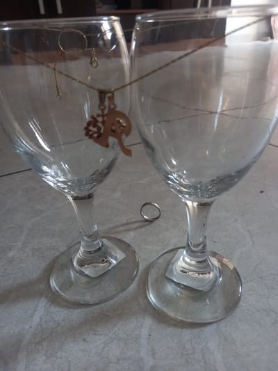 The image depicts two stemmed wine glasses laying horizontally on a tiled surface. Between them sits a silver ring. One of the glasses contains a gold-colored necklace and a separate gold-colored pendant. The photograph appears to be taken indoors. There is no evidence to suggest this is located in Trujillo, Peru, or any specific location.
