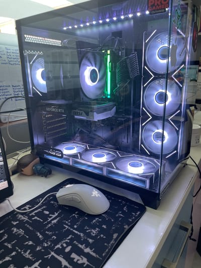 The image shows a gaming computer case with illuminated fans and internal components. A white Razer mouse rests on a black mousepad with a white gun pattern. A small figurine of a person is positioned near the computer case. The computer case is on a white desk, and behind it, a whiteboard with handwritten notes is visible. The scene appears to be in an indoor environment, possibly a room or an office, with no specific indicators pointing to the city of Petrich, Bulgaria, or any particular location within it.