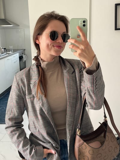 A woman stands indoors, taking a selfie with a light green iPhone 11. She wears round sunglasses, a light brown turtleneck sweater, a plaid blazer in shades of gray and red, and denim jeans, partially visible. A brown shoulder bag with a repeating "G" pattern rests over her blazer. Her brown hair is styled in two braids. The background includes a white wall, a black framed object, and a glimpse of a kitchen with white cabinets and dark flooring, likely located in a residential or commercial space. While the location could be Kranj, Slovenia, there is no definitive visual information within the image to verify it directly.