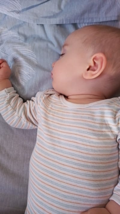 The image is a close-up, top-down view of a baby sleeping on a light blue, patterned sheet. The baby is lying on their right side, facing left, with their head resting on the sheet. They are wearing a long-sleeved onesie with thin horizontal stripes of white, light blue, and light orange. The baby's left arm is bent at the elbow and their hand is resting on the sheet. The right side of the baby's face, ear, and neck are visible. The background consists of crumpled light blue fabric, possibly a blanket or duvet, with a subtle leaf-like pattern in a lighter shade of blue. There are no specific landmarks or indications of the city of Altdorf bei Nürnberg, Germany.