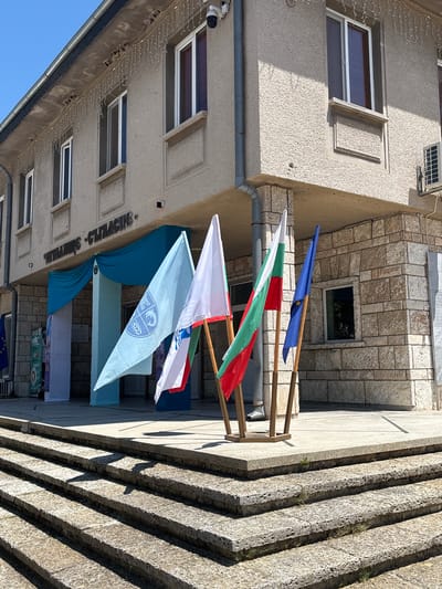 The image depicts the entrance to a building in Kavarna, Bulgaria, likely a municipal or cultural center, as indicated by the Cyrillic inscription above the entrance which reads "ЧИТАЛИЩЕ - СЪГЛАСИЕ" (Chitalishte - Saglasie, meaning "Reading Room - Harmony"). The building has a light-colored stucco facade with stone accents around the lower level and entrance. Several flags are displayed prominently on wooden stands in front of the building. These include the Bulgarian flag (white, green, red), the European Union flag (blue with yellow stars), and another flag with a light blue background and a circular emblem featuring stylized figures and waves. The entrance is adorned with blue drapery. Steps made of rough-hewn stone lead up to the building's entrance. The sky is clear and blue, suggesting a sunny day. The overall scene is festive, possibly in anticipation of or during a public event.