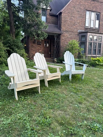 Three Adirondack chairs are positioned on a grassy lawn in front of a two-story brick house. The house has a dark roof and multiple windows, including a bay window on the right side. The address "18908" is visible on a metal grate by the front entrance. The chairs are made of light-colored material, with two being a pale cream color and one on the right appearing to be a very light blue or gray. The grass is green but somewhat unkempt, with some patches of dandelions and other weeds. Lush green trees and bushes surround the house and lawn. The scene appears to be a residential setting in Shaker Heights, Ohio, United States of America, likely in the late afternoon or early evening given the lighting. No people are present in the image.