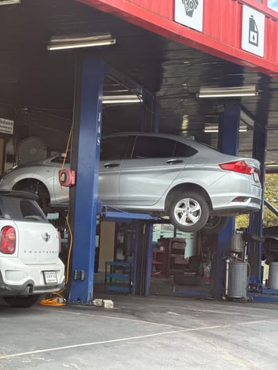 The image depicts a car repair shop in Nonthaburi City Municipality, Thailand. Two cars are visible: a white Mini Cooper Countryman is parked on the ground in the foreground, and a silver sedan is elevated on a hydraulic lift in the background. The repair shop features blue hydraulic lift pillars, fluorescent lighting, and red corrugated metal siding on the exterior. Various tools and equipment, including a red coiled hose and a gray oil drain can, are present in the scene. The ground is paved with asphalt. The overall environment suggests an active automotive service facility.