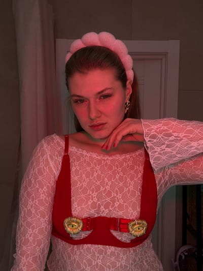 A young woman lies on a surface, angled to the left. She wears a white lace top with flared sleeves, a plush pink headband, and ornate gold earrings. A red, structured harness with decorative gold buckles adorns her chest. Her left hand gently touches her face, drawing attention to her lips and eyes. The scene is bathed in a warm, reddish light. A white wall and part of a window frame are visible in the background.