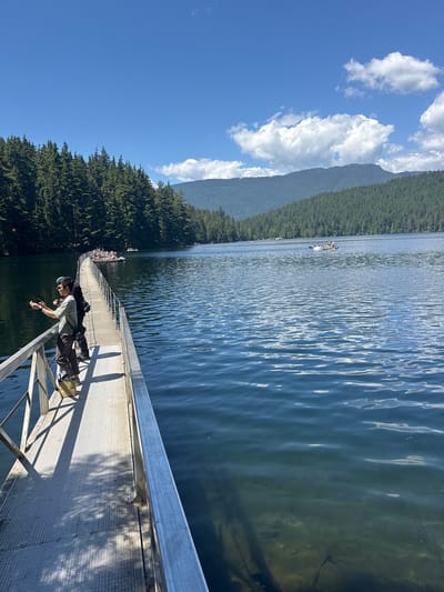 The image depicts a scenic view of a lake on a sunny day, likely at Buntzen Lake in Port Moody, Canada. In the foreground, a person stands on a metal boardwalk that extends over the water. They are holding a fishing rod, suggesting they are engaged in fishing. The boardwalk has a railing and the surface is a textured metal grate. Further down the boardwalk, another person is partially visible. The lake itself is calm with gentle ripples reflecting the blue sky and scattered white clouds. On the far side of the lake, a dense forest of evergreen trees covers the rolling hills, which rise to meet the mountains in the distance. Several small boats, some carrying groups of people, are scattered across the water, indicating recreational activity. The overall atmosphere is peaceful and natural.