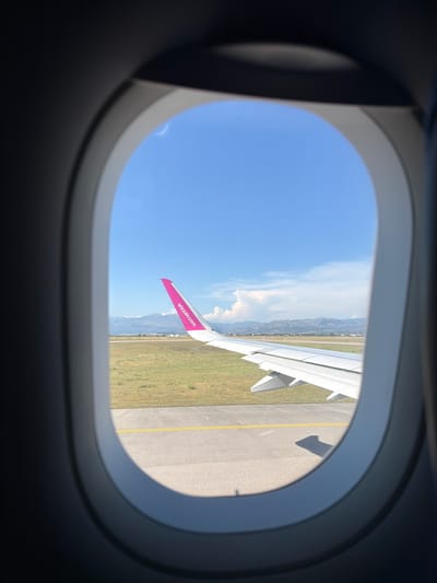 The image shows an airplane window view of an airport runway under a clear blue sky with scattered clouds. The right side of the image features a portion of the airplane's wing, which is white with a bright pink wingtip adorned with the "wizzair.com" logo. Beyond the runway, there is green grass and a distant landscape of mountains and some buildings. The perspective suggests the airplane is either on the ground at the airport or has just taken off/is about to land. The location is specified as Donja Cijevna, Montenegro.