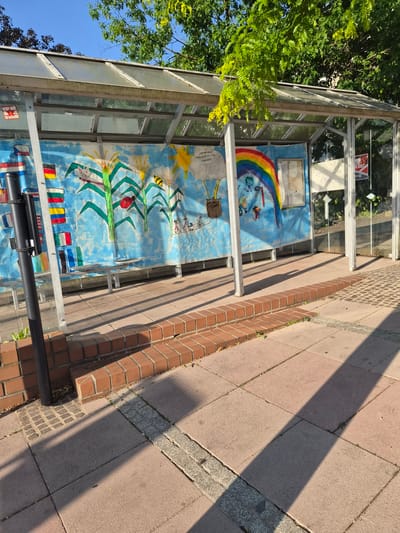 The image shows a bus stop shelter with a mural painted on the back wall. The mural depicts a blue sky with clouds, corn stalks, a sun, a rainbow, and various figures including insects, a ladybug, children on bicycles, and people playing basketball. The mural also features flags of different countries on the left side.
The bus stop shelter has a metal frame with a transparent roof and walls. There are benches inside the shelter. The ground in front of the shelter is paved with reddish-brown tiles. A low brick wall runs along the edge of the paved area.
The image was taken in Sanem, Luxembourg, on a sunny day. The sun is casting long shadows on the ground. There are trees visible in the background. There are no people visible in the image.