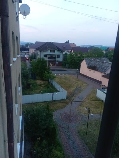 The image shows an exterior view of a street in Tiachiv, Ukraine, likely taken from a window of a multi-story building. The scene is set during dusk or early morning, with a soft, overcast sky. Several buildings are visible, including a light green multi-story building with balconies and windows, and a peach-colored single-story building with a dark-tiled roof. A metal fence runs along a grassy area. A paved pathway with a circular mosaic pattern made of bricks is prominent in the foreground, with a street lamp with two bulbs situated nearby. In the distance, more buildings and trees are visible, as well as rolling hills. A red car is seen on the street, and a group of people, appearing to be children, are standing near the peach-colored building. A satellite dish is attached to the side of the building in the foreground. The overall atmosphere is calm and quiet.