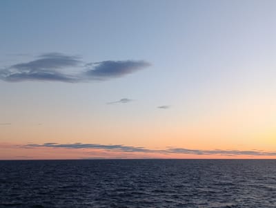 The image depicts a serene seascape at dusk. The sky transitions from a pale blue at the top to soft hues of peach and orange near the horizon. Wispy clouds are scattered across the sky, with larger, darker formations concentrated in the upper left quadrant. A thin band of clouds stretches across the mid-horizon, tinged with the colors of sunset. The sea below is a deep, dark blue, with gentle ripples creating a textured surface. The horizon line is sharp and clear, separating the sky from the water. There are no people, structures, or identifiable landmarks visible in the scene, suggesting an open, natural environment. The overall impression is one of tranquility and natural beauty, characteristic of coastal Finland, and specifically could be a view from the shores of Larsmo, Finland, known for its extensive coastline and archipelago.