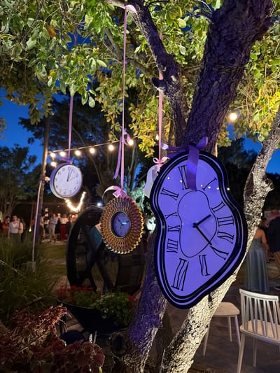 The image shows an outdoor event space at night in Nir Eliyahu, Israel. Clocks are hanging from tree branches by pink ribbons. The clocks are of various styles: a melting clock reminiscent of Salvador Dali's work, a sunburst clock, and a standard round clock. String lights are also strung through the trees. People are present in the background, standing and talking. Some white chairs are also visible.