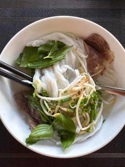 A close-up overhead shot features a bowl of pho, a Vietnamese noodle soup, resting on a dark textured placemat. The bowl is white and filled with a light-colored broth. Prominently visible are wide, flat white rice noodles, thin slices of cooked beef, and a generous garnish of fresh green herbs, including basil leaves and possibly cilantro. Bean sprouts and chopped green onions are also scattered throughout the dish. A pair of black chopsticks rests on the edge of the bowl, partially submerged in the broth, as is a rose gold-colored spoon. The scene is devoid of any people, indicating an unattended dish. The setting does not provide specific visual cues to pinpoint its location within Quy Nhon, Vietnam.
