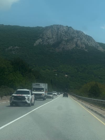 The image shows a line of vehicles on a winding road adjacent to a mountain covered in dense green vegetation. The lead vehicle is a white car with the word "TAXI" visible on its side. The other vehicles in the line include a flatbed truck and several other cars, all traveling in the same direction. The road is marked with a white line separating the lanes and a yellow line along the edge. The environment includes a large rocky mountain on one side of the road and a lush green hillside on the other. Based on the location information provided, the scene is likely in Орлиновский округ, Ukraine.