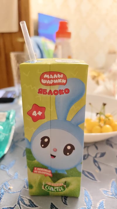 A juice box with a cartoon bunny character is shown, with a straw inserted. The juice box is green and yellow, with text in Russian. The bunny is blue and white, with large eyes. The juice box has a red star with "4+" inside. The background is blurred and appears to be an indoor setting, possibly a dining table, with a bowl of yellow fruit in the background. The scene is likely in Занарацкі сельскі Савет, Belarus, as indicated by the text on the juice box.