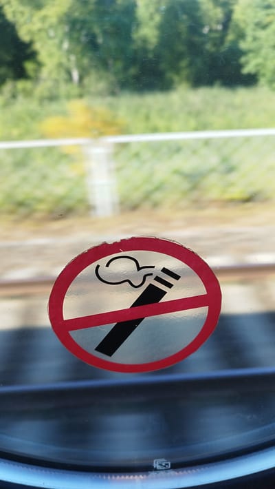 A circular "no smoking" sign with a red border is adhered to a glass surface. The sign depicts a stylized cigarette with smoke and is crossed out by a red diagonal line. The background is blurred and shows greenery and trees, suggesting an outdoor environment viewed through a window. The lower portion of the image shows a reflective surface, possibly a train window, with horizontal lines visible. There are no people or explicit actions depicted in the image.