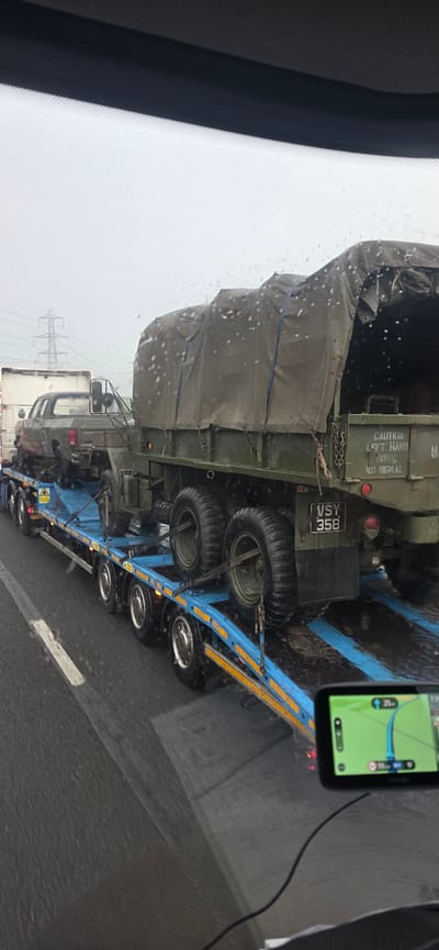 A trailer carrying a military truck and a pickup truck is visible on a wet road, likely in Uttlesford, United Kingdom, given the presence of power lines in the background consistent with the rural landscape of the region. The military truck, green and covered with a tarp, is the primary subject. A pickup truck, dark colored, is loaded onto the front of the trailer, positioned in front of the military truck. The trailer itself is blue, with multiple wheels and reflective yellow markings along its sides. The road surface is wet, indicating rain, and water droplets are visible on the windshield from which the photo is taken. A GPS device is mounted in the foreground, displaying a navigation map. The overall scene suggests a vehicle transport on a highway during overcast weather.