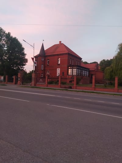 The image shows a red brick building with a conical tower and a red tiled roof, located in Gusev, Russia. The building is surrounded by a low brick fence with metal railings. A street with white lines runs in front of the building. Trees and a street lamp are also visible in the scene. The sky is a soft pink and blue, suggesting it is either dawn or dusk. There are no people visible in the image.