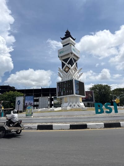 In Lambaro, Indonesia, a modern monument with the "BSI" logo stands tall against a partly cloudy sky. The monument features multiple tiers, with the "BSI" branding prominently displayed on its upper sections. A large digital screen is integrated into the monument's base, showcasing a complex array of colorful pixels, suggesting it may be displaying data or advertisements. To the right of the monument, large, teal-colored "BSI" letters are positioned on the ground, further reinforcing the branding. In the background, a partially constructed building, likely a commercial or public structure, is visible, with its skeletal framework exposed. Several billboards are also present, one featuring an advertisement with two figures in uniform and text that appears to read "SELAMAT TAHUN BARU." To the left of the monument, a motorcycle with a small trailer attached is moving away from the camera, driven by a person wearing a helmet and a patterned shirt. The road in the foreground is asphalt, marked with a yellow line and a black and white striped curb. Lush green trees are visible behind the billboards and the construction site, indicating a well-developed urban environment. The overall scene depicts a busy public space with a focus on commercial branding and urban development.