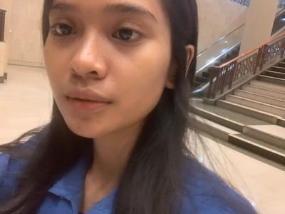 This media file contains a rotated selfie of a young adult female, captured indoors. The subject has long, dark hair that partially frames her face and wears a blue garment. Her dark eyes gaze directly towards the camera with a neutral or slightly serious expression. A notable detail on her face is a slight redness or chapping on her lower lip.

The background suggests a spacious, multi-level indoor public area, potentially a shopping mall, hotel, or large commercial building. Architectural features visible include light-colored walls and ceiling, with prominent horizontal beams. On what appears to be an upper level or balcony, there is an ornate railing with intricate, possibly carved, patterns, indicating a decorative architectural style. The scene is illuminated by artificial indoor lighting, making the time of day undeterminable. No specific activity beyond taking the selfie is depicted, and no visible text is present in the image. The general appearance of the individual and the setting is consistent with the provided location context of Samarinda, Indonesia.