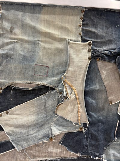 The image is a close-up shot of denim patchwork art, featuring various shades of blue denim, pieced together with metal studs along some of the edges. The denim pieces show wear and tear, with frayed edges and visible seams. A portion of a zipper is visible near the top center of the image. The background consists of more denim patchwork, creating a textured and layered effect. The scene is likely an indoor setting, given the controlled lighting. There's no indication of Spreitenbach, Switzerland, or any specific location. No people are visible.
