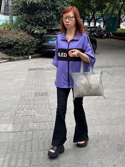 A street-level shot shows a woman standing on a sidewalk in 龙溪街道, China. She has red hair and wears glasses, a purple shirt with black accents, black pants, and black sandals. She carries a semi-transparent gray shopping bag. To her left is a parked car, vegetation, and street signs. In the background, there are motorbikes and buildings partially visible. The sidewalk is paved with gray tiles, and the area appears to be a mix of residential and commercial.
