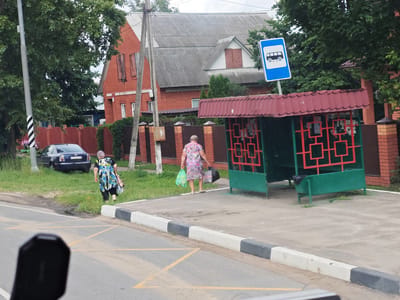 The image depicts a street scene in Kabunovo, Russia. A bus stop structure, painted green with a red metal roof and decorative red lattice work, is situated on the right side of the frame. A blue bus stop sign is mounted on a pole to the left of the bus stop. Two women are visible; one is walking towards the bus stop, carrying two plastic bags, one green and one black. She is wearing a colorful floral dress and dark leggings. The second woman, further to the left and walking away from the camera, is wearing a blue and white patterned dress and dark leggings. A dark-colored sedan is parked on the grassy area to the left of the women. In the background, a red brick house with a gray corrugated roof stands, partially obscured by trees. The street in the foreground has yellow crosswalk markings. A utility pole with black and white diagonal stripes is on the far left. The environment appears to be a residential or semi-rural area with vegetation and trees present.