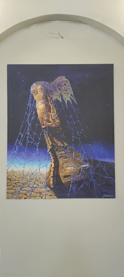 The image shows a painting of a golden, humanoid figure entangled in blue spiderwebs against a dark, starry sky. The figure appears to be a statue or an ancient artifact, with intricate details on its torso and head, and a blue gemstone embedded in its chest. Below the figure, a cracked, arid landscape stretches towards a horizon with a gradient of blue and yellow. The painting is signed "Yelena" in the bottom right corner. The painting is hung on a plain white wall, beneath a curved architectural element. There are no people visible in the image. The scene depicted in the painting is not specific to Voskevaz, Armenia, or any identifiable real-world location.