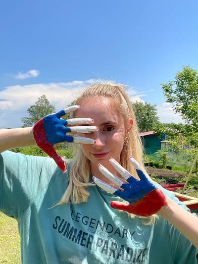 A young woman with blonde hair pulled into a ponytail is shown outdoors, with a blue sky and scattered clouds above. She is wearing a light teal t-shirt with the text "LEGENDARY SUMMER PARADISE RELAX & SURF" on it. Her face is partially obscured by her left hand, which is held up to her face. The woman's hand is painted with red and blue stripes, and her fingernails are painted white. Her right hand is also visible, with fingers painted white. The background consists of greenery, trees, and what appears to be a small rural settlement with houses and garden beds. The lighting suggests a sunny day. The setting is likely outdoors in a rural or suburban area, potentially the city of Yasnoe, Russia, given the context.
