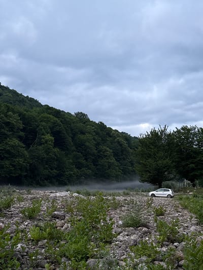 This is a vertically oriented, slightly hazy photograph taken from a roadside perspective. The scene depicts a lush, forested mountainside on the left, sloping upwards towards a cloudy sky. A narrow, misty stream or waterfall appears to cascade down a portion of the mountainside. On the right side of the frame, a rocky embankment is visible, covered in patches of green foliage. A white car is parked on the unpaved roadside area between the trees and the rocky embankment. The time of day appears to be daytime, possibly overcast given the light and cloud cover. The location is Abgara, Abkhazia.
