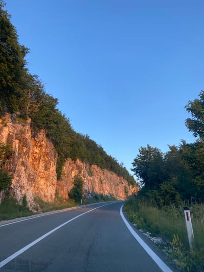 This image shows a winding asphalt road with white lane markings, bordered by a steep, rocky cliff face on the left and trees and foliage on the right. The cliff is illuminated by warm, golden sunlight, suggesting it is either sunrise or sunset. Lush green trees and vegetation cover the top of the cliff and the right side of the road. A white road marker with a red stripe is visible on the right edge of the road. The sky is a clear, bright blue. The scene appears to be a natural, mountainous area, likely outside of a densely populated urban environment. The image does not depict any people or specific landmarks that would place it within the city of Praga, Montenegro.