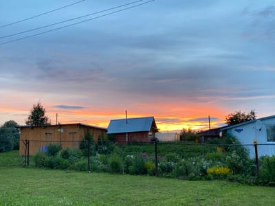 This media file depicts a serene rural scene in Баклуши, Russia, at dusk. The sky is ablaze with the warm hues of sunset, transitioning from vibrant oranges and yellows near the horizon to soft blues and grays higher up, with wispy clouds catching the fading light. Three distinct structures are visible: a long, light-brown wooden outbuilding with a corrugated metal roof on the left, a small, dark-wood log cabin with a metal roof and a chimney in the center, and a light-blue house with a satellite dish on the right. A chain-link fence runs across the foreground, separating a neatly mowed green lawn from a lush garden filled with a variety of flowering plants in shades of pink, white, and yellow, as well as dense green foliage. Behind the garden, a greenhouse is partially visible. The overall atmosphere is peaceful and idyllic, evoking a sense of calm and rural tranquility. The time of day is clearly indicated by the sunset and the soft, diffused lighting. Visible text on the light-blue house reads "ул. ЗАРЕЧНАЯ 21А".