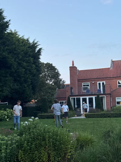 This image captures a residential garden scene in Bubwith, United Kingdom, at dusk. Four males are present on a manicured green lawn. To the left, a large, dense evergreen tree dominates the mid-ground. The foreground features an array of green foliage and white flowers. In the background, a multi-story brick house with a red tiled roof is prominent, featuring a tall brick chimney topped with a reflective cap, a balcony with a dark railing and two blue chairs, and a ground-floor conservatory with white-framed glass panels.
On the lawn, one male wearing a light-colored t-shirt and blue jeans stands towards the left. In the central area, two males are in motion: one, viewed from behind in a light shirt and dark trousers, holds a yellow, stick-like object; the other, in a white t-shirt and dark shorts, moves forward. A fourth male, partially obscured behind the central figures near the house, also holds a yellow, stick-like object. A dark spherical object, consistent with a ball, rests on the grass near the central figures. The collective arrangement of individuals and objects suggests an outdoor game in progress.