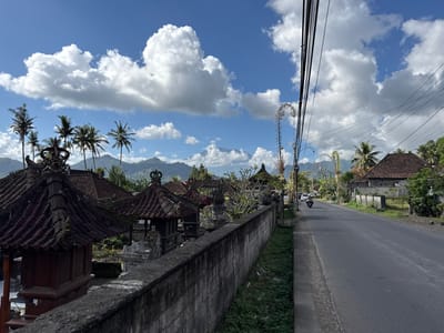 In Sidemen, Indonesia, a sunlit day is depicted with a bright blue sky dotted with voluminous white clouds. A paved road stretches into the distance on the right side of the frame, bordered by a narrow grassy verge and a low, weathered stone wall. Beyond the wall, traditional Balinese structures with dark, multi-tiered tiled roofs and ornate carvings are visible, interspersed with lush green foliage and palm trees. In the background, verdant, rolling hills rise to meet the sky. On the road, a person rides a motorcycle, heading away from the viewer. Overhead, a dense network of black electrical wires crisscrosses the sky, supported by poles. The overall impression is one of a tranquil, rural Indonesian setting under a clear sky.