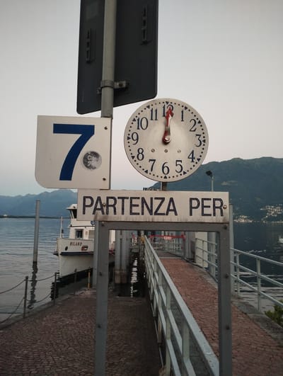 The image depicts a pier or dock area next to a body of water, likely Lake Maggiore, in Muralto, Switzerland. A white passenger boat, identified by the name "MILANO" on its side, is docked on the left. Overhead, a clock face shows the time as approximately 12:05. A sign above the boat reads "PARTENZA PER," meaning "Departure for." Another sign to the left displays a large blue numeral "7" within a white square. The walkway leading from the shore is paved with cobblestones and bordered by a metal railing. In the background, hills covered with trees and houses are visible, with a hazy sky overhead. The overall atmosphere is that of a transportation hub, possibly a ferry terminal. There are no people visible in the immediate scene.