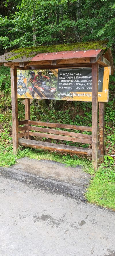 A wooden, open-sided shelter with a gabled roof and an integrated bench stands beside a paved road in a verdant, mountainous environment in Ribaritsa, Bulgaria. The structure is constructed from dark, rough-hewn timber posts and beams. Its roof is covered with reddish-brown corrugated material, heavily encrusted with green moss. Beneath the roof, a horizontal banner is affixed, advertising ATV trips.

The banner features a graphic of two individuals on a red ATV navigating a muddy track, with a "TV TRIP" logo including "RENT & ADVENTURE" and "ETROPOLE." To the right, Cyrillic text translates to: "ATV TRIP FOR RENT IN THE MOUNTAINS + INSTRUCTION, EQUIPMENT, MOUNTAIN GUIDE, FOR GROUPS OF 1 TO 10 PEOPLE." A website address, "WWW.ATVFORESTRIP.COM," is visible at the bottom of the banner. The bench consists of a thick, rounded log serving as the seat and three horizontal wooden planks forming the backrest.

The ground immediately around the structure is covered in green grass and small plants. In the foreground, a grey asphalt road with visible aggregate and darker wet patches extends across the bottom of the frame. The background is dominated by a dense, lush forest of deciduous trees with vibrant green foliage, indicating a natural, unpopulated setting. No individuals or active events are present in the scene.