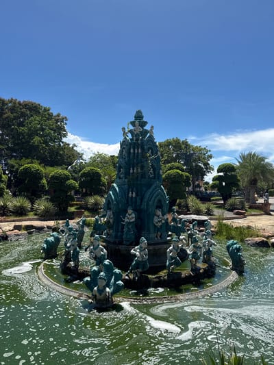A tall, ornate, greenish-blue sculpture/fountain structure resides in a body of greenish water, with a foam-like substance floating on its surface. The sculpture appears to be multi-tiered and adorned with human figures. Around the water feature, there are landscaped areas with trees and plants. The sky is blue with some clouds. It is located in the city of Bang Pu Subdistrict Municipality, Thailand.