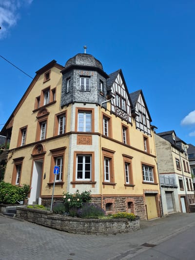 This detailed image depicts a sunny street scene in Traben-Trarbach, Germany, centered on a historic corner building.

The primary subject is a multi-story building featuring a light yellow stucco facade with contrasting brown window frames and trim. A prominent grey slate-tiled octagonal turret defines the corner, topped with a finial. The turret's upper section displays decorative slate work including the numbers "1908" in contrasting lighter slate. Below the turret, on the corner of the facade, an ornate carved relief contains the initials "F.D." and the date "1908". The right side of the building transitions into traditional German half-timbered construction on its upper stories, characterized by dark wooden beams and white infill. The ground floor of this building, and extending to the right, features a dark, rough-cut stone foundation. A small, light yellow garage door is visible on the ground level of the main building's right side.

To the right of the main building, the yellow stucco and half-timbered architectural style continues on an adjacent structure. Further down the street, another building with a lighter-toned, textured facade, a metal-railed balcony, and a ground-level garage door is partially visible.

The street and sidewalk in the foreground are paved with grey interlocking pavers. A raised stone retaining wall and planter in front of the main corner building contain various plants, including green bushes, pink roses, and purple lavender. A blue rectangular street sign with a white horizontal bar, indicating a dead-end street, is prominently displayed on a pole in front of the building. A metal drain cover is visible in the darker asphalt section of the road to the right.

The sky is a clear, vibrant blue with minimal scattered white clouds, indicating bright, sunny weather. No people are present in the scene.