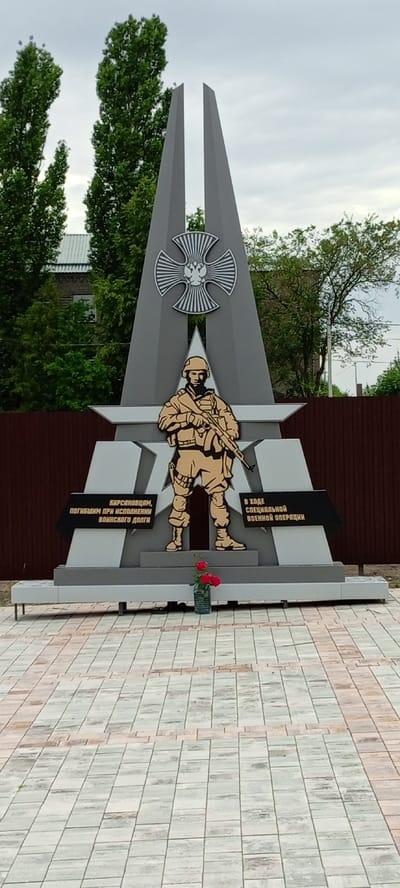 The image shows a monument in the city of Kirsanov, Russia. The monument is a tall, grey structure with a stylized cross at the top, featuring the Russian coat of arms. In the center, a silhouette of a soldier in uniform, holding a rifle, is depicted. Below the soldier, there are two black plaques with inscriptions in Russian. The left plaque reads "КИРСАНОВЦАМ, ПОГИБШИМ ПРИ ИСПОЛНЕНИИ ВОИНСКОГО ДОЛГА" (To Kirsanovites, who perished while fulfilling their military duty). The right plaque reads "В ХОДЕ СПЕЦИАЛЬНОЙ ВОЕННОЙ ОПЕРАЦИИ" (During the special military operation). In front of the monument, on a tiled pavement, there is a vase with red roses. The monument is situated in front of a brown wooden fence, with trees and buildings visible in the background. The sky is overcast.
