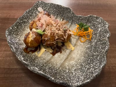 A close-up shot features a serving of six golden-brown takoyaki (octopus balls) presented on a speckled grey and white ceramic plate with an irregular, wavy edge. Each takoyaki is topped with a generous amount of thin, light brown katsuobushi (bonito flakes) and small pieces of green onion. Dark brown takoyaki sauce and creamy yellow Japanese mayonnaise are drizzled over the items and onto the plate. A garnish of fine julienned orange carrots and a sprig of green herb is positioned on the upper right side of the plate. The plate rests on a smooth, dark reddish-brown wooden surface. No people, specific actions, or interactions are visible. The precise location within the city of Bath, United Kingdom, cannot be determined from the visual information provided.