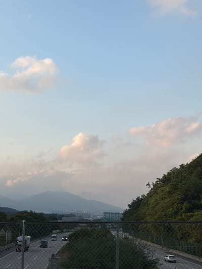 This is a wide-angle, outdoor, ground-level shot of a highway that appears to be located in or near Pocheon-si, South Korea, during daylight hours. The sky is mostly clear with scattered, wispy clouds, tinged with pink and white, suggesting it might be late afternoon or early evening. In the foreground, a chain-link fence runs horizontally across the bottom of the image.
Beyond the fence, the highway curves to the left and recedes into the distance. Several vehicles are visible on the road, including a white truck in the left lane, a white car in the right lane, and a few other cars further down the road. The highway is bordered by lush green trees and vegetation on the right side, which forms a dense hillside. On the left side of the highway, there are some barriers and more greenery.
In the background, a hazy mountain range dominates the horizon, with a cityscape visible in the valley between the mountains and the highway. The buildings in the cityscape are densely packed and appear to be residential or commercial structures. The overall impression is a blend of urban development and natural landscape.