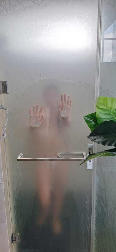 A person's hands are pressed against a frosted glass shower door, obscuring their body. The hands are facing outward, with fingers splayed. One hand has a ring on the ring finger. Water droplets are visible on the glass. To the right of the shower door, a large green plant is partially in view. A silver handle is attached to the shower door, and a sign that reads "PUSH" is visible on the right side of the door. The background appears to be a bathroom. The image does not provide information to determine a specific location in Payangan, Indonesia.