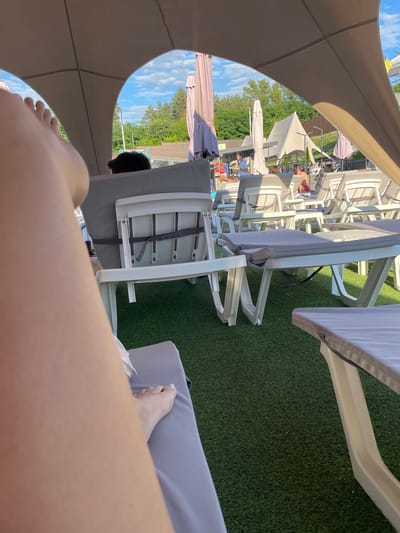 The image depicts an outdoor lounge area covered by a large, light-colored canopy, likely at a resort or pool facility in Zazymia, Ukraine. In the foreground, a person's leg and foot are visible, resting on a gray lounge chair. The foot is adorned with white nail polish. Beyond the foreground, multiple white plastic lounge chairs with gray cushions are arranged on an artificial green turf surface. Some chairs are occupied by individuals, including a man in swim trunks and several others partially visible. In the background, trees and a cloudy blue sky are visible. Several large, light-colored umbrellas and a tent-like structure are also present. The overall scene suggests a leisure setting, possibly during daytime given the lighting.