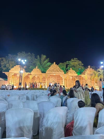 Here is a description of the image:

The image captures a night scene in Nashik, India, during what appears to be an outdoor event, potentially a wedding or large celebration. The backdrop consists of an elaborate, ornate structure with intricate carvings, illuminated by bright lights. Trees and other vegetation are present, adding to the outdoor setting. Rows of chairs covered in white fabric extend to the right of the frame, suggesting seating arrangements for attendees. People dressed in various traditional Indian attires can be seen mingling and standing in front of the illuminated structure. The left side of the image is dominated by a dark night sky, contrasting with the brightly lit event area. The overall scene suggests a festive and celebratory occasion.