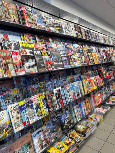 This is a vertical, eye-level, medium shot of a magazine display rack. The rack is filled with numerous magazines of varying sizes and colors, creating a vibrant and busy visual. The magazines are neatly arranged on shelves, with some standing upright and others leaning against each other. The subjects of the magazines are diverse, including cars, motorcycles, homes and gardens, lifestyle, and sports. Many of the magazine covers feature bold text, images of vehicles, people, and scenic landscapes.

The lighting suggests it is daytime, with artificial light illuminating the magazines from above. The setting appears to be a retail environment, likely a newsstand, bookstore, or supermarket, given the organized display of numerous publications. The overall impression is one of abundance and variety, showcasing the wide range of interests and information available through print media. The text visible on the magazines includes titles and captions in various languages, such as Dutch, French, and possibly German. Examples of visible text include "HOUSE & GARDEN," "Auto Classics," "Porsche Magazine," "LIVING," "WIELER," and titles related to cycling events like the "Tour de France." The sheer volume of magazines and the detailed covers suggest a large selection is available to potential customers.