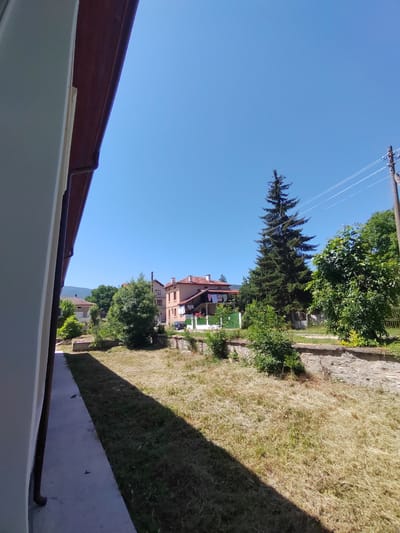 The image depicts a sunny daytime scene in Rakitovo, Bulgaria, likely in a residential area. A light-colored building or wall is in the foreground on the left, with a dark brown gutter. Beyond this, a dry, grassy yard with scattered small bushes stretches towards a stone retaining wall. Several houses with red-tiled roofs are visible in the middle ground, along with a tall evergreen tree. Clothes are hanging on a line outside one of the houses. A black car is parked on a road to the right of the houses. In the background, a power pole with wires and a line of trees are visible against a clear blue sky. There are no people or discernible actions taking place in the image.